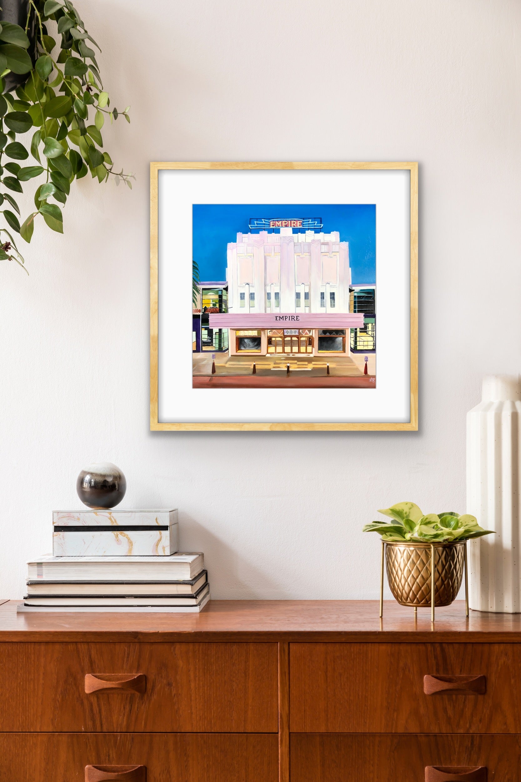 Framed artwork of the Empire Theatre in Toowoomba on a wall above a wooden cabinet with decorative items. A limited edition print by Leonie McIntosh